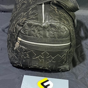 K&ouml;nnte beinhalten: Schwarze Leder-Duffel-Tasche mit einem Kreuzmuster, das &uuml;ber die Oberfl&auml;che gepr&auml;gt ist. Die Tasche hat einen silbernen Rei&szlig;verschluss und eine kleine Rei&szlig;verschlusstasche auf der Vorderseite. Sie hat einen Griff oben und einen Schultergurt.