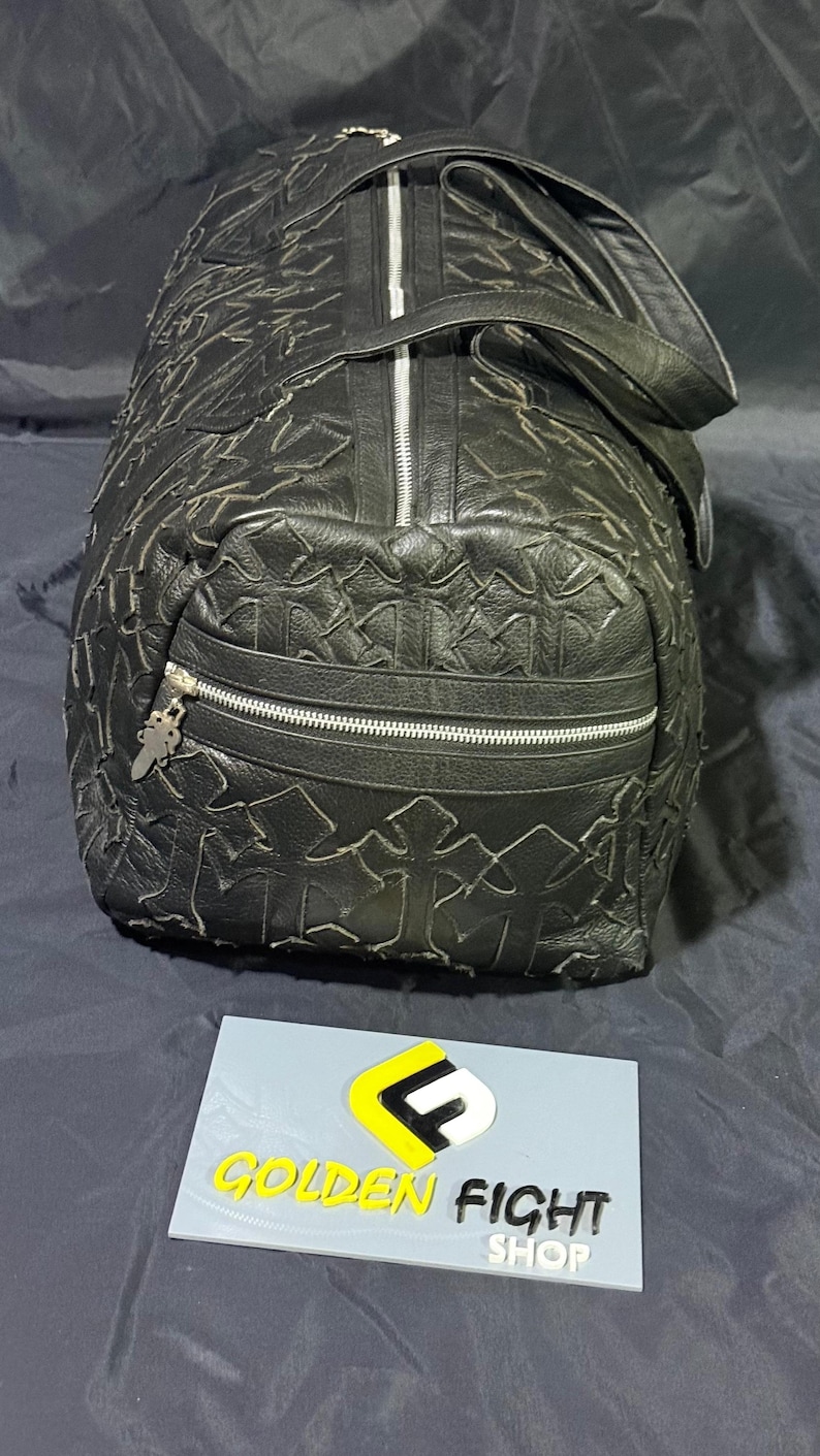 K&ouml;nnte beinhalten: Schwarze Leder-Duffel-Tasche mit einem Kreuzmuster, das &uuml;ber die Oberfl&auml;che gepr&auml;gt ist. Die Tasche hat einen silbernen Rei&szlig;verschluss und eine kleine Rei&szlig;verschlusstasche auf der Vorderseite. Sie hat einen Griff oben und einen Schultergurt.