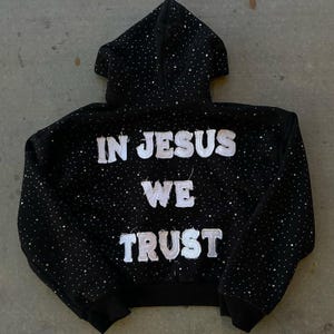 Pu&ograve; includere: Felpa con cappuccio nera con cappuccio e maniche lunghe. La felpa &egrave; impreziosita da piccoli accenti scintillanti e presenta la scritta "IN JESUS WE TRUST" in bianco, con lettere strutturate sul retro.