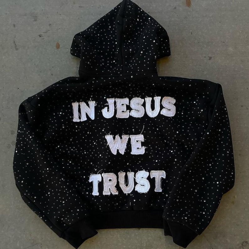 Pu&ograve; includere: Felpa con cappuccio nera con cappuccio e maniche lunghe. La felpa &egrave; impreziosita da piccoli accenti scintillanti e presenta la scritta "IN JESUS WE TRUST" in bianco, con lettere strutturate sul retro.