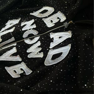 Pu&ograve; includere: Felpa con cappuccio nera con cerniera, con dettagli di strass bianchi e le parole "ALIVE NOW DEAD" in lettere bianche, usurate e cucite. La felpa ha una cerniera argentata e un cappuccio.