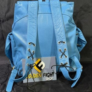 May include: A light blue leather backpack with adjustable straps and side pockets. The backpack features black lacing details and a top handle. The text "GOLDEN FIGHT SHOP" is visible on a sign attached to the backpack.