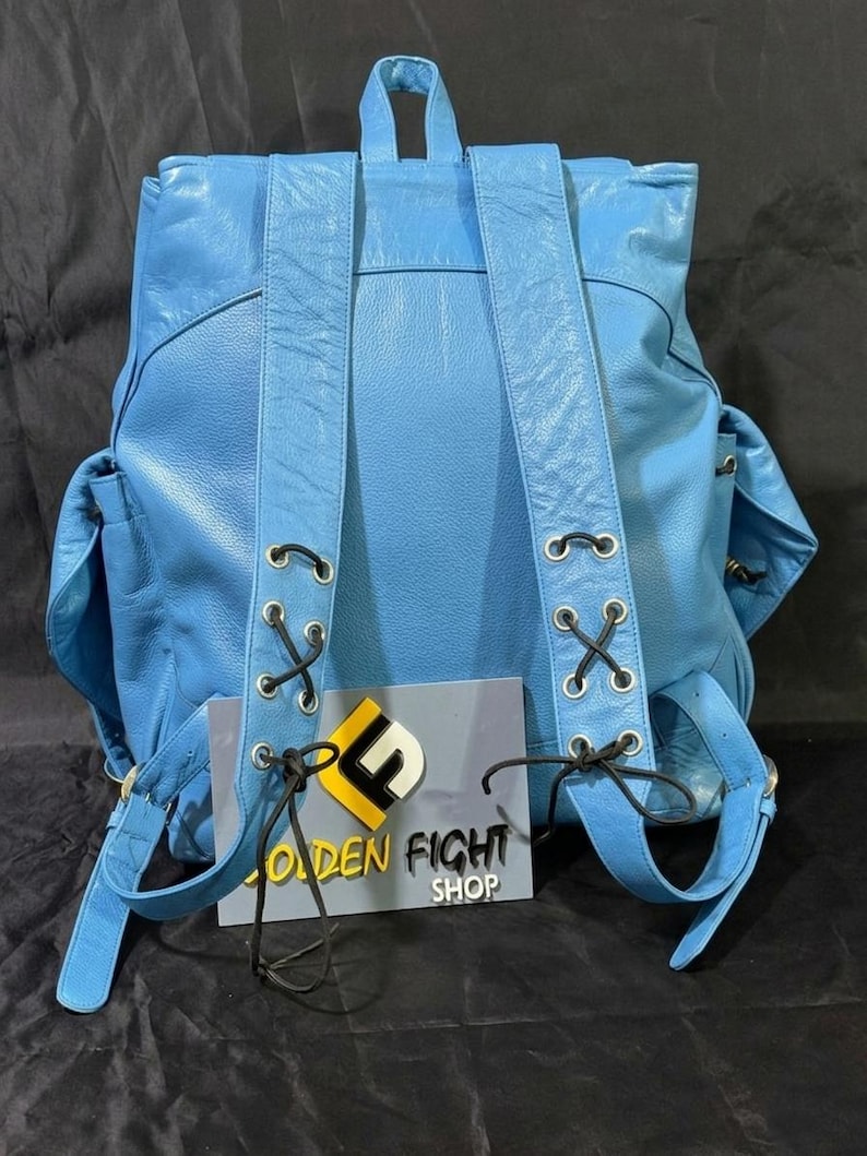 May include: A light blue leather backpack with adjustable straps and side pockets. The backpack features black lacing details and a top handle. The text "GOLDEN FIGHT SHOP" is visible on a sign attached to the backpack.