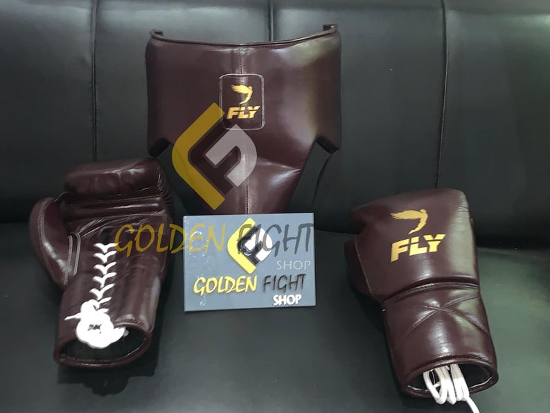 May include: A boxing set in brown leather, including gloves, headgear, and a protective cup. The gloves have white laces and the brand "FLY" is visible. A sign reads "GOLDEN FIGHT SHOP".
