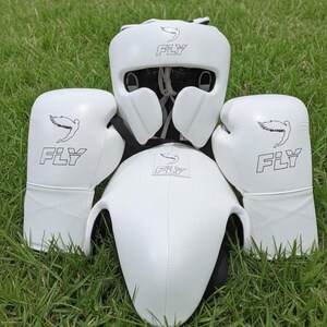 Puede incluir: Un conjunto de boxeo blanco completo, que incluye protector de cabeza, protector de pecho y guantes de boxeo. Cada pieza presenta la palabra "FLY" y un gráfico de pájaro. El conjunto está sobre hierba verde.