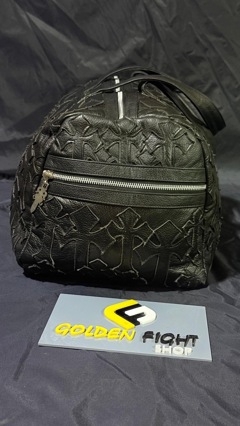 K&ouml;nnte beinhalten: Schwarze Leder-Duffel-Tasche mit Kreuzmuster. Die Tasche hat eine Rei&szlig;verschlusstasche auf der Vorderseite und einen silbernen Rei&szlig;verschluss. Die Tasche hat einen oberen Griff und einen silbernen Anh&auml;nger am Rei&szlig;verschluss. Die Tasche befindet sich auf einer dunklen Oberfl&auml;che.