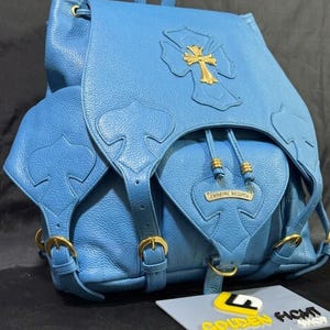 May include: A light blue leather backpack with a gold cross emblem and gold hardware. The backpack has a drawstring closure, adjustable straps, and multiple pockets. The brand name "CHROME HEARTS" is visible on the front pocket.
