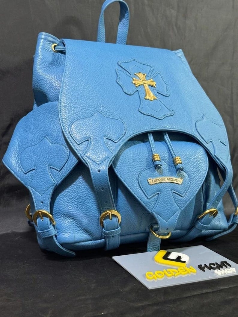 May include: A light blue leather backpack with a gold cross emblem and gold hardware. The backpack has a drawstring closure, adjustable straps, and multiple pockets. The brand name "CHROME HEARTS" is visible on the front pocket.