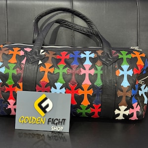Könnte beinhalten: Schwarze Leder-Duffel-Tasche mit bunten Kreuzapplikationen. Die Tasche hat einen Reißverschluss, zwei Griffe und eine kleine Seitentasche. Ein Schild vor der Tasche lautet "GOLDEN FIGHT SHOP".