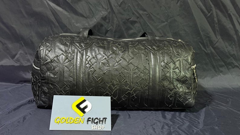 K&ouml;nnte beinhalten: Schwarze Leder-Reisetasche mit sich wiederholendem Kreuzmuster. Die Tasche hat zwei Griffe und einen Rei&szlig;verschluss. Das Logo von Golden Fight Shop ist im Vordergrund.