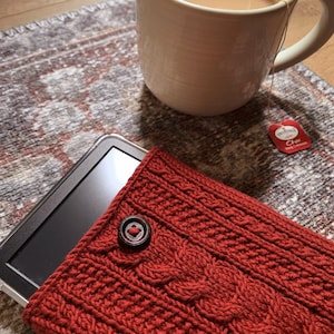Peut inclure: Une housse de tablette en tricot rouge avec une fermeture à bouton. La housse est sur un tapis avec une tasse de thé et un sachet de thé.