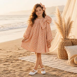 May include: A long-sleeved dress featuring a white and orange gingham check pattern, a large bow detail on the left shoulder, and a matching hair bow. The dress has a loose, flowing silhouette with gathered sleeves and a round neckline.