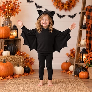 May include: A black long-sleeve hooded sweatshirt featuring attached bat wings extending from the sides and small pointed ears on the hood, paired with matching black pants. The garment is designed with a soft texture and a solid black color.