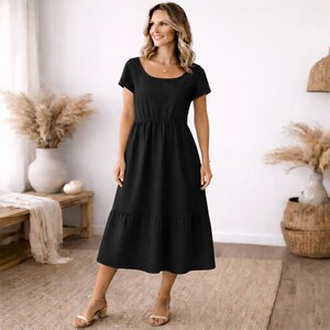 Peut inclure: Une robe midi noire avec un col rond et des manches courtes. La robe pr&eacute;sente une taille ajust&eacute;e et une jupe &agrave; volants. Le mod&egrave;le porte des sandales beiges. L'arri&egrave;re-plan comprend un banc en bois, un vase avec des plantes s&eacute;ch&eacute;es et un panier tress&eacute;.