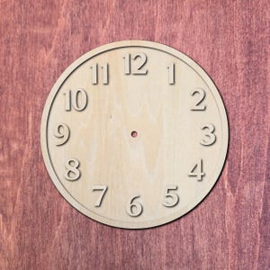 May include: A circular light-colored wooden clock face featuring raised numerals from one to twelve arranged in a standard clock pattern. The center of the disc has a small circular hole for a clock mechanism, and the wood grain is visible.
