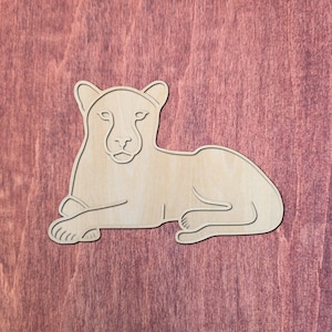 Puede incluir: Un recorte de madera de color claro con la forma de una pantera en reposo. La figura presenta líneas grabadas que definen los rasgos faciales, las orejas y la postura corporal del felino, con una superficie lisa y veta de madera natural.