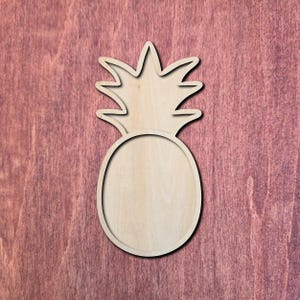 May include: A wooden pineapple cutout with a detailed outline. The pineapple has a textured, natural wood grain appearance. The design includes the spiky crown and the oval body of the fruit. The cutout is set against a reddish-brown background.