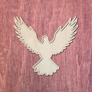 May include: A flat, light-colored wooden cutout in the shape of a bird with wings spread wide in flight. The silhouette features detailed feather edges along the wings and a fan-shaped tail, showcasing the natural grain of the wood material.