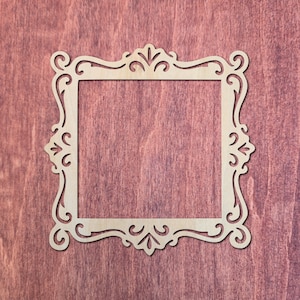 May include: A square wooden frame cutout featuring intricate scrollwork and filigree patterns along all four sides. The light-colored wood has a smooth, flat surface with a symmetrical design that includes curved lines and pointed decorative elements.