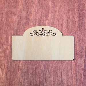 May include: A light-coloured wooden sign with a decorative cutout design at the top. The sign has a rectangular base with a curved top, and the cutout features a floral and swirl pattern. The background is a reddish-brown wood.