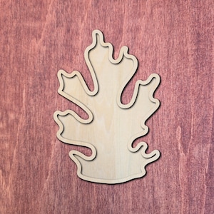 May include: A light-coloured, unfinished wooden cutout shaped like a stylised oak leaf. The piece features a smooth, flat surface with a distinct, continuous outline that follows the irregular, lobed edges of the leaf form, creating a simple silhouette.