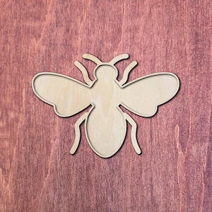 Puede incluir: Un recorte de madera de color claro con forma de abeja. La silueta del insecto presenta una cabeza redondeada, un tórax central, un abdomen más grande y dos alas simétricas que se extienden desde los lados, con dos antenas arriba.