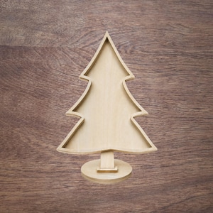 May include: A light-coloured wooden Christmas tree-shaped tray with a raised border and a flat base. The object features a smooth, natural wood grain finish and a symmetrical silhouette, standing upright on a small, circular pedestal support.