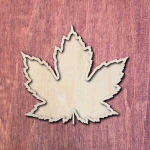 May include: A flat, light-coloured wooden cutout in the shape of a maple leaf. The leaf features a detailed, jagged edge profile with a central stem at the bottom. The surface of the wood shows a natural, light beige grain pattern throughout.