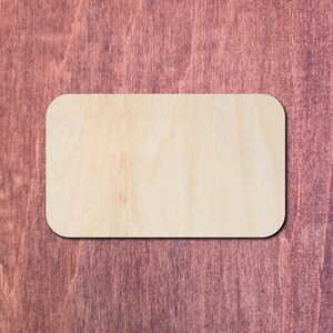 May include: A rectangular piece of light-coloured, unfinished wood with rounded corners. The surface displays a natural wood grain pattern with visible light tan and beige tones, featuring a smooth, flat texture across the entire front face.