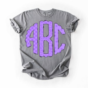 May include: A gray t-shirt featuring a purple monogram design. The monogram includes the letters "ABC" in a decorative, scalloped border. The letters are filled with a checkered and leopard print pattern. The shirt is made of a soft material.