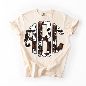 May include: Cream-colored t-shirt featuring a large monogram design with the letters ABC. The monogram has a brown and white cowhide pattern, with a scalloped border. The shirt has short sleeves and a crew neck.