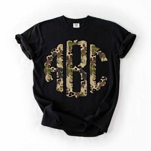 May include: Black t-shirt featuring a large, camouflage-patterned monogram of the letters ABC. The monogram is centered on the shirt and stands out against the dark background. The shirt has short sleeves and a crew neck.
