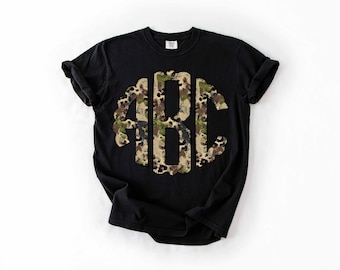May include: Black t-shirt featuring a large, camouflage-patterned monogram of the letters ABC. The monogram is centered on the shirt and stands out against the dark background. The shirt has short sleeves and a crew neck.
