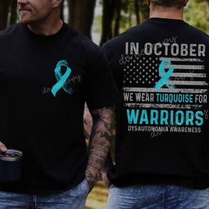 Op de afbeelding: Zwart T-shirt met een turquoise lint op de voor- en achterkant. De achterkant toont een Amerikaans vlagontwerp en de tekst "IN OCTOBER WE WEAR TURQUOISE FOR WARRIORS DYSYTONOMIA AWARENESS". Het shirt bevordert bewustwording van dysautonomie.