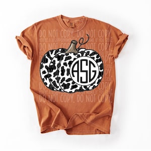 May include: An orange t-shirt with a black and white cow print pumpkin design and the initials "ASG" in a circular monogram. The shirt has short sleeves and a round neckline. The words "DO NOT COPY" are printed on the shirt.