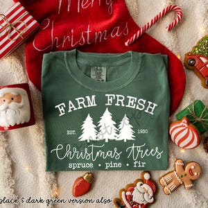 May include: A dark green t-shirt with the text "FARM FRESH Christmas Trees" and three white Christmas trees. The shirt is surrounded by Christmas decorations, including a red stocking, candy cane, and gingerbread cookies.