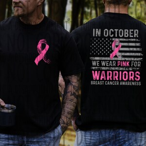 May include: Black t-shirt with a pink ribbon graphic. The back displays an American flag design with the text: "In October We Wear Pink For Warriors Breast Cancer Awareness."