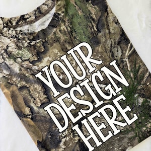 May include: A camouflage-patterned t-shirt with the text "YOUR DESIGN HERE" in a bold, white font. The shirt features a crew neck and a woodland camouflage design in shades of brown, green, and tan.