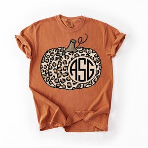 Puede incluir: Camiseta naranja quemado con un diseño de calabaza con estampado de leopardo y las iniciales "ASG" en un monograma circular. La calabaza tiene un tallo marrón y está centrada en la camiseta. La camiseta tiene mangas enrolladas.