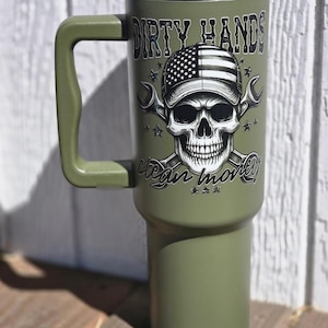 May include: Olive green tumbler with a handle, lid, and straw. The tumbler features a graphic of a skull wearing a cap with an American flag design, crossed wrenches, and the text "DIRTY HANDS CLEAN MONEY."
