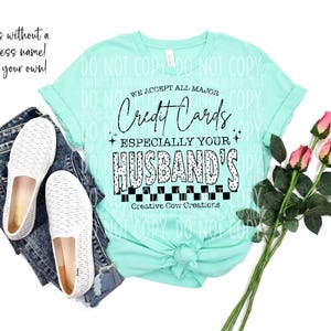 May include: A light turquoise t-shirt with the text "WE ACCEPT ALL MAJOR Credit Cards ESPECIALLY YOUR HUSBAND'S". The shirt is paired with blue jeans, white shoes, and pink roses. The text "Comes without a business name! Add your own!" is also visible.