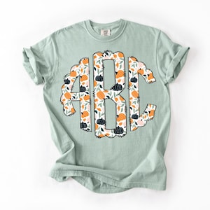 Op de afbeelding: Een lichtgroen t-shirt met een monogramontwerp van de letters "ABC". Het monogram is gevuld met een patroon van oranje en zwarte pompoenen, groene bladeren en witte bloemen. Het shirt heeft opgerolde mouwen.