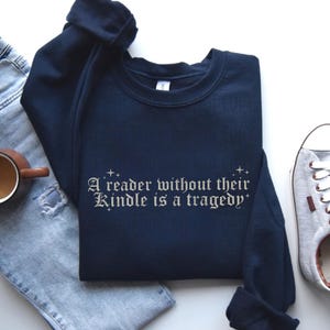 May include: Navy blue sweatshirt with the text "A reader without their Kindle is a tragedy" in a decorative font. The sweatshirt is paired with light blue jeans, a brown mug, and gray sneakers.