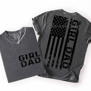 May include: Two charcoal gray t-shirts. One shirt has the words "GIRL DAD" printed in black. The other shirt has a distressed American flag design with the words "GIRL DAD" printed vertically. A silver chain necklace is on the first shirt.