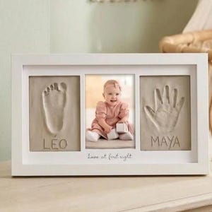 May include: White framed baby keepsake with a photo and imprints of a foot and hand. The photo shows a baby in a pink outfit. Text includes the names "Léo" and "Maya" and the phrase "Love at first sight."