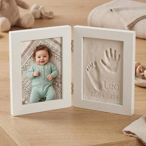 Peut inclure: Cadre photo blanc à charnière. Un côté affiche une photo de bébé, l'autre une empreinte de main et de pied. L'empreinte est nommée "Léo" et datée du 22.04.2024. Le bébé sur la photo porte une combinaison vert clair.