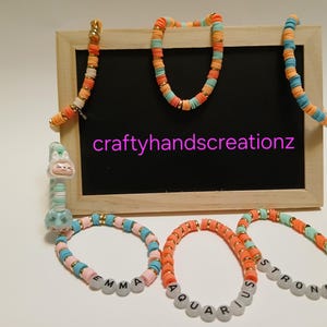May include: A collection of colorful beaded jewelry, including necklaces and bracelets. The bracelets feature letter beads spelling out names and words like "AQUARIUS" and "STRONG". The background includes a small chalkboard with the text "craftyhandscreationz".