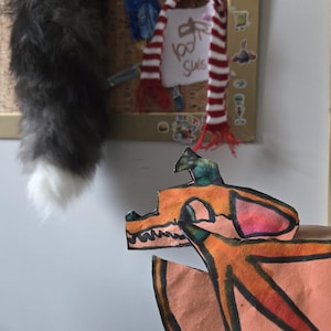 May include: A corkboard with a cat tail, a red and white striped scarf with the text "100 Subs", and a paper cutout of a dragon with orange, pink, and green colors.