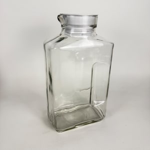 May include: A clear glass pitcher with a rectangular body and a wide, integrated handle on the side. The vessel features a short, cylindrical neck topped with a circular, metallic-colored lid that fits securely over the opening of the container.