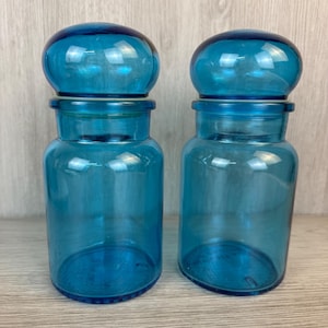 May include: Two matching cylindrical glass jars in a vibrant translucent blue color. Each jar features a smooth, rounded, dome-shaped lid that sits securely on top. The glass is clear, allowing light to pass through, highlighting the deep blue hue.